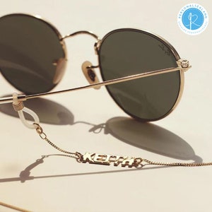 Op de afbeelding: Ronde, goudkleurige zonnebril met een gouden ketting die aan de pootjes is bevestigd. De ketting heeft een gouden naamplaatje met de tekst "KEITH".