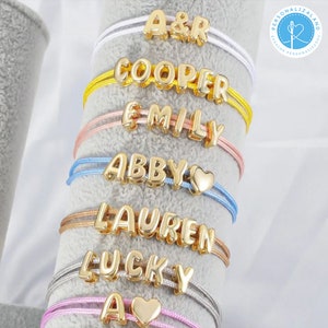 Op de afbeelding: Gouden naamarmbanden met namen zoals "COOPER" en "EMILY" worden getoond. Elke armband heeft gouden letterkralen op gekleurde koorden, waaronder wit, geel, roze, blauw en grijs. Sommige armbanden hebben hartvormige bedels.