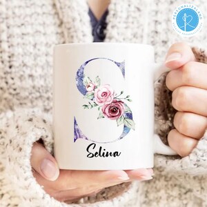 May include: White ceramic mug featuring a purple and blue watercolour letter "S" adorned with pink and white flowers. The name "Selina" is written beneath the letter.