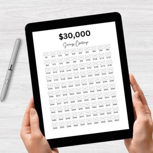 May include: A black and white digital tablet displaying a savings challenge tracker with a grid of 100 envelopes. Each envelope has a dollar amount printed on it, starting at $6 and increasing by $2 up to $500. The text "$30,000 Savings Challenge" is at the top of the tablet.