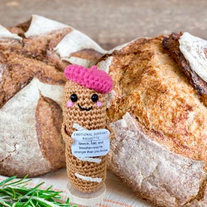 May include: A crocheted baguette doll wearing a pink beret and a smiling face, holding a sign that reads "EMOTIONAL SUPPORT BAGUETTE". Two loaves of crusty bread are in the background, with rosemary sprigs in the foreground.