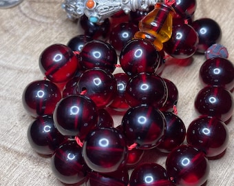 May include: A red amber prayer beads necklace with a silver tassel and a silver and turquoise bead accent. The beads are strung on a red cord.