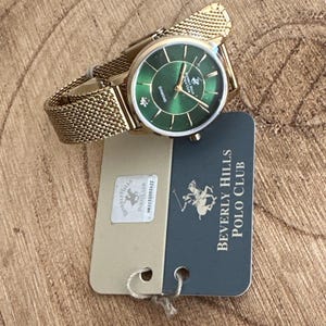 May include: A gold-toned wristwatch with a green dial and a mesh band. The watch face features the text "Diamond" and the Beverly Hills Polo Club logo. A tag with the brand name is attached.