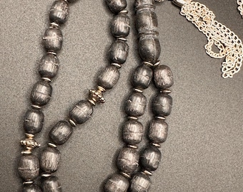 Puede incluir: Un collar de cuentas gris oscuro con cuentas en forma de barril y detalles plateados. El collar tiene una cadena plateada y una borla decorativa. Las cuentas tienen una superficie texturizada y están unidas con conectores plateados.