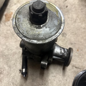 May include: A used, metallic grey automotive component with a black cap and various fittings. The part shows signs of wear and age. The design includes a circular top and a cylindrical body.