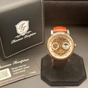 Pode incluir: Um relógio Thomas Tompion dourado com uma pulseira de couro castanho e um mostrador esqueleto, exibido em um suporte preto. O relógio está ao lado de uma caixa preta com o logotipo Thomas Tompion e um cartão de garantia.