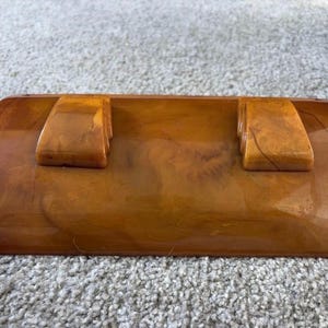 Puede incluir: Un objeto semicilíndrico de color ámbar con un acabado liso y brillante y una textura veteada. Presenta dos bloques rectangulares elevados situados en la superficie superior, cada uno con detalles laterales escalonados.