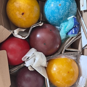 May include: A collection of six spherical bowling balls in various colours including yellow, blue, red, and dark brown, packed inside a cardboard box with protective dividers. Some balls feature a textured finish, while others are smooth and solid.