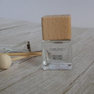 May include: A clear glass car diffuser with a wooden cap, labelled "Lychee Blush" by Golden Serenity Candle Co. Includes a wooden ball and diffuser sticks. The diffuser is 15ml.