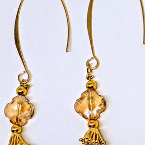 May include: A pair of gold-colored earrings with a hook design. Each earring features a cluster of small gold beads, a four-leaf clover-shaped amber-colored crystal, and a gold tassel. The earrings are displayed against a white background.