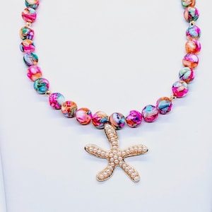 Könnte beinhalten: Eine farbenfrohe Perlenkette mit einem goldfarbenen Seestern-Anhänger. Die Kette hat runde Perlen in Rosa, Orange, Grün und Blau. Der Seestern-Anhänger ist mit kleinen, weißen, perlenartigen Perlen verziert.