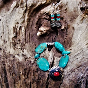May include: Turquoise and red stone earrings and bracelet set with silver accents. The jewellery is displayed on a piece of weathered wood.