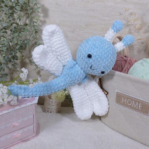 Puede incluir: Un peluche de libélula tejido a ganchillo con hilo suave y texturizado en azul claro y blanco. La libélula presenta una cabeza redonda con ojos de seguridad negros, dos antenas con segmentos azules y blancos y cuatro alas blancas redondeadas.