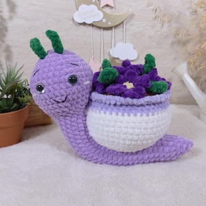 Peut inclure: Un escargot en crochet peluche avec un corps violet clair, deux antennes vertes et des yeux de sécurité noirs. L'escargot possède une coquille blanche servant de pot, remplie de fleurs en crochet violet foncé et de petites feuilles vertes.