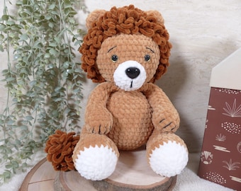 May include: A plush lion toy crafted from soft brown yarn with a textured, curly dark brown mane. The lion features a white muzzle, a black plastic nose, embroidered black eyes, and white paws. A fluffy brown tail is visible to the left of the body.