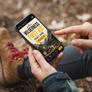May include: A smartphone displaying a digital Wilderness First Aid Pocket Guide. The screen shows text and an image of an olive green first aid kit, a compass, and a metal mug against a mountain backdrop. The interface includes navigation icons.