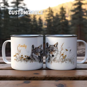 May include: Two white enamel camping mugs with a black rim and handle. The mug on the left has the name "Emily" printed on it. Both mugs feature a wolf and wolf pup design with a watercolour style and a brown, white, and grey colour scheme.