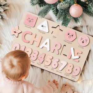 May include: A wooden puzzle board with pink and white pieces featuring the name "CHARLIE" and numbers 1-9. The board also includes shapes with faces, such as a square, triangle, star, hexagon, and circle. A baby is playing with the puzzle on a white fluffy surface.