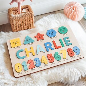 May include: A wooden puzzle board with colourful shapes, numbers, and letters. The board features a square, triangle, star, hexagon, and circle with happy faces. The name "CHARLIE" is spelled out in large letters, and the numbers 0 through 9 are displayed below.