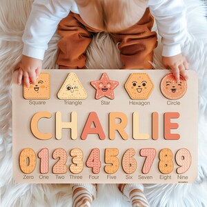 May include: A wooden educational toy with a light brown background. The toy features a variety of shapes, including a square, triangle, star, hexagon, and circle. Each shape has a smiling face and is labelled with its name. The toy also includes the name "CHARLIE" in large, colourful letters and the numbers 0 through 9 with a patterned design.