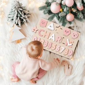 May include: Wooden puzzle with the name "CHARLIE" and numbers 0-9 in pink and white. The puzzle includes shapes like squares, triangles, and stars. The background features a Christmas tree and a child wearing pink clothing.