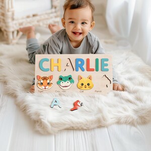 May include: A wooden personalized puzzle board with the name "CHARLIE" spelled out in colorful letters. The board features animal faces, including a tiger, crocodile, cat, and parrot, with corresponding labels below each animal. The puzzle pieces are missing from the board, and a few pieces are scattered on a white fluffy surface.