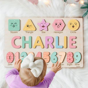 May include: A wooden puzzle board with colourful shapes, numbers, and letters. The board features a square, triangle, star, hexagon, and circle, each with a cute face. The numbers zero through nine are also included, along with the name "CHARLIE" in large, colourful letters.
