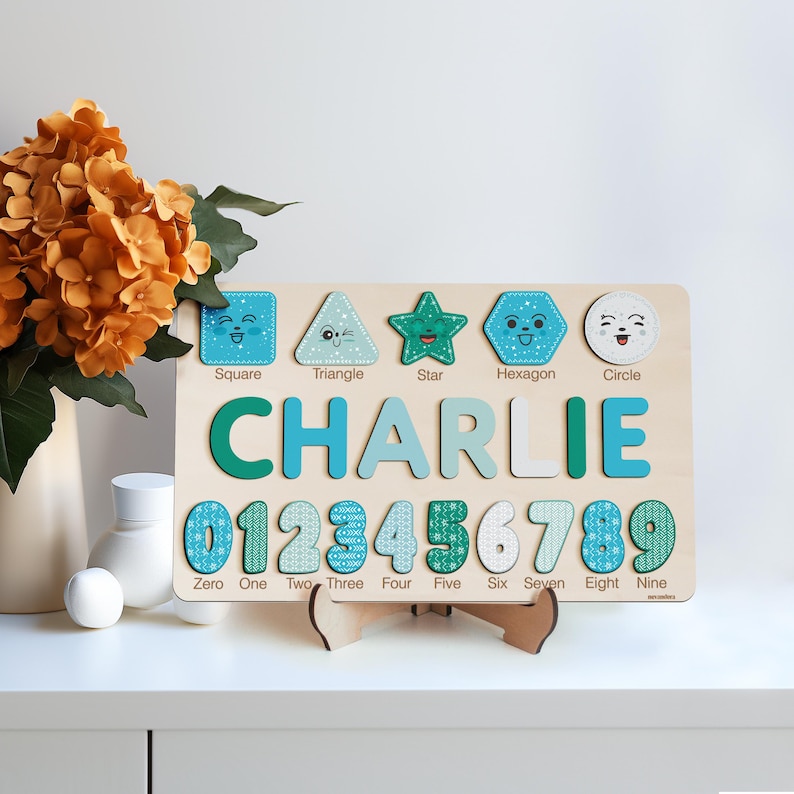 May include: A wooden educational toy with a blue and green colour scheme. The toy features a child's name, "CHARLIE", along with numbers 0-9, and shapes with corresponding names: square, triangle, star, hexagon, and circle.