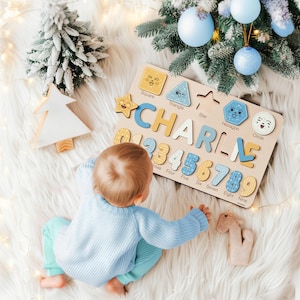 May include: Wooden puzzle board with the name "CHARLIE" spelled out in individual letters. The board also features numbers and shape cutouts. The puzzle is on a white fluffy surface, with a baby in a blue sweater and pants.