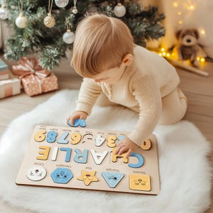 May include: A wooden puzzle with the name "CHARLIE" and numbers 1-9. The puzzle also includes shapes like a circle, hexagon, star, triangle, and square. The child is playing with the puzzle on a white fluffy rug.