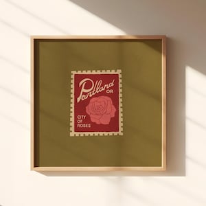 May include: Square framed art print featuring a vintage-style postage stamp design. The stamp, in red, displays the word "Portland OR" and a rose illustration, with "City of Roses" below. The background is olive green.