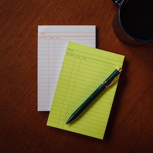 May include: Two notepads, one white and one yellow-green, with grid lines and the words "TITLE", "DUE DATE", "DONE", and "TASK" printed at the top. A dark green pen with gold accents rests on the yellow-green notepad. A dark blue mug is in the background.