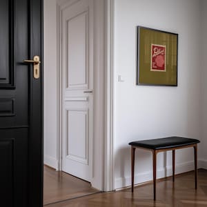 May include: A black leather bench with wooden legs sits against a white wall. A framed print with a red stamp design hangs above the bench. The image is taken in a hallway with a black door and a white door.