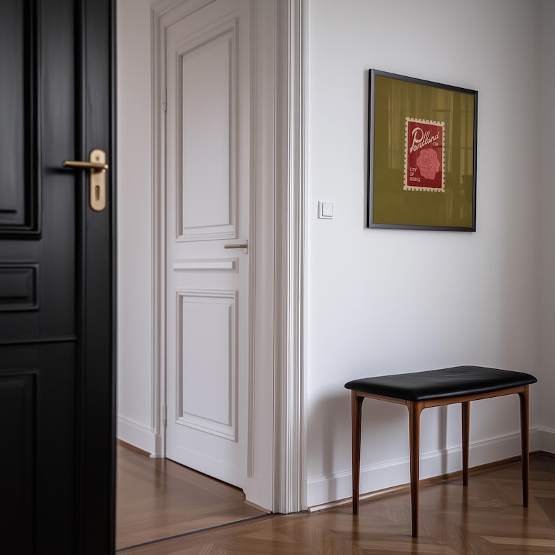 May include: A black leather bench with wooden legs sits against a white wall. A framed print with a red stamp design hangs above the bench. The image is taken in a hallway with a black door and a white door.