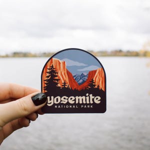 May include: A sticker featuring a scenic illustration of Yosemite National Park. The design includes red rock formations, pine trees, and the text "yosemite NATIONAL PARK" in white. The sticker has a dark blue and brown border.