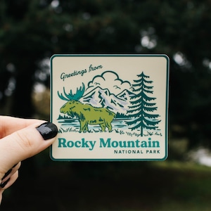 May include: Square sticker with a green and white illustration of a moose, mountains, and a pine tree. The text "Greetings from Rocky Mountain National Park" is printed on the sticker.