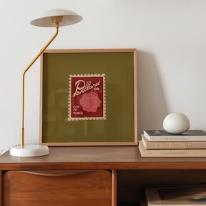 May include: A framed print featuring a red postage stamp design with the words "Portland OR City of Roses" and a rose illustration. A white and gold lamp and a stack of books are on a wooden surface.