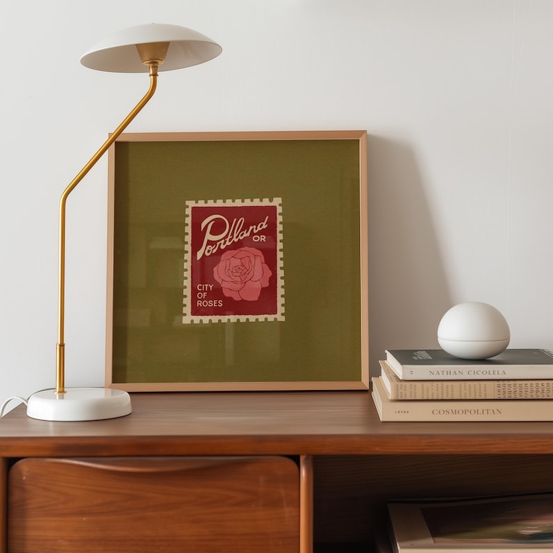 May include: A framed print featuring a red postage stamp design with the words "Portland OR City of Roses" and a rose illustration. A white and gold lamp and a stack of books are on a wooden surface.