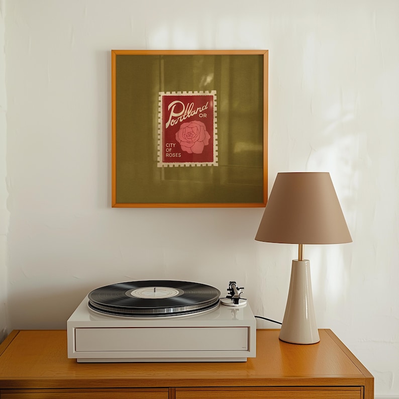 May include: A framed print of a Portland, Oregon stamp with a rose design hangs above a white record player and a beige lamp with a conical shade. The lamp sits on a wooden surface.