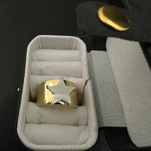May include: A gold ring with a white star-shaped stone in a black ring box with white velvet lining.