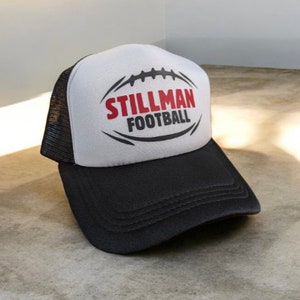 May include: A white and black trucker hat with a black mesh back. The front of the hat has a black and white football graphic with the text "Stillman Football" in red and black.