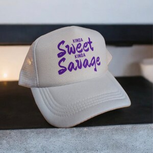 May include: A white trucker hat with a purple embroidered design that says "Kinda Sweet Kinda Savage".