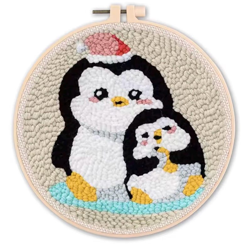 May include: A handmade, round embroidery hoop featuring two cartoon penguins. The larger penguin wears a red and white Santa hat. The penguins are black, white, and yellow, set against a beige background and a light blue base.