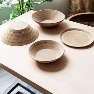 May include: A collection of light brown, circular ceramic items including a stack of five graduated bowls, one single bowl with visible horizontal ridges, and one flat circular plate, all featuring a matte, unglazed, earthy texture.