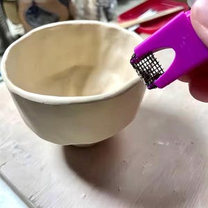 May include: A cream-colored ceramic bowl with a rounded shape. A purple tool with a mesh screen is held near the bowl. The bowl sits on a light-colored surface, likely a work table. The image suggests a pottery or ceramics project.