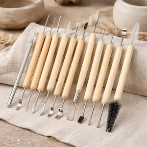 May include: A collection of pottery tools featuring wooden handles and stainless steel heads. The set includes tools with different shapes for sculpting and detailing, such as loops, needles, and a brush. The tools are displayed on a beige cloth.