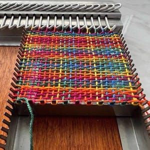 Può includere: Un tessuto multicolore in fase di lavorazione, creato su un telaio in metallo. La sezione tessuta mostra una vibrante gamma di colori, tra cui rosso, arancione, giallo, verde, blu e viola. Un filo verde è attaccato alla tessitura. Il telaio è in metallo.