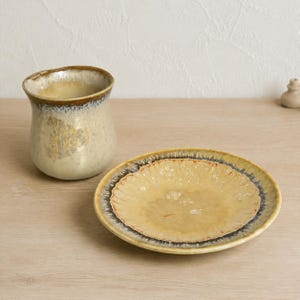May include: A ceramic cup and a matching saucer featuring a reactive glaze finish. The set displays a creamy beige center that transitions into a dark, mottled brown rim with a glossy texture. The cup has a wide, slightly flared opening.