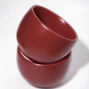 May include: Two maroon ceramic bowls, one stacked on top of the other. The bowls have a smooth, glossy finish and a rounded shape. They are likely for serving food or as decorative items. The bowls are a deep red colour.