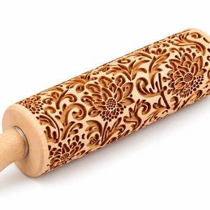 Könnte beinhalten: Ein helles Nudelholz aus Holz mit einem tief eingravierten Blumenmuster auf dem zentralen Zylinder. Das Design besteht aus komplexen, sich wiederholenden chrysanthemenartigen Blüten und geschwungenen Rankenmotiven, die in das Holz geschnitzt sind.